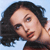 Pixelated close-up of a person with dark wavy hair against a light blue background, showing partial face and shoulder.