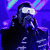Pixelated image of a person in dark clothing holding a microphone under blue lighting, next to a cartoon bear wearing white glasses with striped lenses.