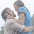 Two people embrace outdoors; one wears a white shirt, the other blue. The background is light with blurred greenery.