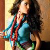 A woman with long dark hair wears a blue sleeveless top and red beaded necklace, standing against a light brown wall with her left hand on her hip.