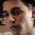 Pixelated close-up of a person's face with dark hair, visible eyes, nose, and mouth, set against a light background with indistinct shapes.