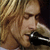 A pixelated image showing a person with long blonde hair singing into a microphone on the left, and bold yellow text reading "KURT LIVES ON!!!" on a black background on the right.