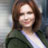 Pixelated static image of a person with shoulder-length brown hair wearing a dark green jacket over a white shirt against a blurred background.