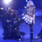 Animated sequence of a performer in plaid skirt and gray top dancing with backup dancers on blue-lit stage, captured from multiple angles.