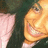 A smiling person with long dark hair and light skin, wearing a pink garment under a dark outer layer.