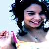 A person with curly dark hair wearing a light-colored top with pink straps and a silver necklace with a blue pendant, smiling with visible teeth against a light background.