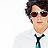 Pixelated static image of a person's face and upper torso in formal attire with dark hair, sunglasses, and a white shirt with black tie against light background.