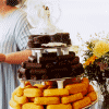A person in a light blue dress pours white liquid from a pitcher onto a three-tiered chocolate cake with white icing, surrounded by orange pastries and yellow flowers.