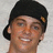 Pixelated close-up of a person with short dark hair wearing a black cap, smiling broadly against a light gray background.