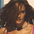 A pixelated close-up of a person with voluminous dark brown hair, wearing a light-colored top with pink straps and a silver necklace, set against a blurred background.