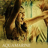 A woman with long brown hair lies on a sandy surface, mouth open in a scream, right arm raised, wearing a light-colored dress with vertical stripes.