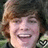 Pixelated close-up of a person with light brown hair and a checkered shirt, smiling with visible teeth against a dark background.