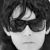 Pixelated grayscale image of a person's face with dark hair, wearing dark sunglasses, and a neutral expression.