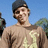 Pixelated image of a person wearing dark cap and light brown shirt with yellow design against blue sky background.