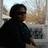 A person with dark hair and sunglasses sits on a porch, wearing a black shirt with green accents against a blurred outdoor background.