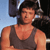 A pixelated image of a person wearing a dark sleeveless shirt, standing against a background with blue sky and dark structures.