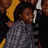 A child in a gray patterned shirt and dark pants stands against a yellow and black background with other figures partially visible.