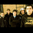 Four pixelated figures in dark clothing stand against a light brown background with visible texture.