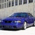 Blue car with dark blue accents parked in front of a light-colored building facade featuring vertical rectangular windows.