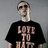 Pixelated static image of a person wearing dark sunglasses and a black T-shirt with large light-colored text on the chest against a gray background.