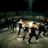 A pixelated group of figures in dark clothing on a textured background with one figure holding a bright light source.