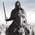 Pixelated grayscale figure in dark hooded cloak holding sword, standing against light gradient background with scattered pixel clusters.