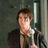 Pixelated image of a person with brown hair wearing a dark jacket over a light shirt, standing against a dark textured background with a vertical light-colored structure.