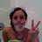 A person with dark hair in a ponytail wearing a brown top and green bottom makes a peace sign against a light gray background.