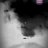 A pixelated grayscale image with a central dark shape against a light background, featuring a small dark spot below the shape and a faint purple pixel cluster in the top-right corner.