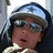 Pixelated close-up of a person wearing a dark helmet with a light blue reflective visor, dark sunglasses, and a light-colored shirt, set against a blurred background.