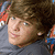 A young person with brown hair and blue eyes wears a gray shirt against an orange-and-blue patterned background.