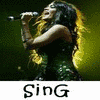 A woman with long dark hair sings into a microphone, wearing a dark sequined dress against a green background with yellow lighting.