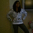 A pixelated figure with long dark hair wears a light gray sweater and black pants, standing in an indoor setting with muted brown tones.