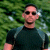 A pixelated image shows a person wearing dark sunglasses and a dark short-sleeve shirt with light blue shoulder accents, standing against a green leafy background.