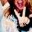 Pixelated image of a person with long brown hair wearing dark clothing, holding up two fingers in front of their face against a light blue background.