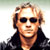 Static pixelated image of a person with light hair wearing dark sunglasses and a black jacket against a gradient background.