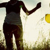 Silhouetted figure in motion wearing striped tank top and dark pants, arms extended, standing in tall grass with bright overexposed background.