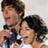 Two people with dark hair stand close together; one holds a microphone, both have open mouths suggesting singing or speaking.