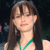 A pixelated portrait of a person with long brown hair and bangs, wearing a green necklace against a dark background.