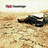Static pixelated image showing a dark aircraft on sandy terrain with a blurred red-and-gray object above.