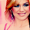 A close-up of a person with light brown hair, blue eyes, and a pink top, wearing long dangling earrings against a pink background.