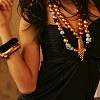 A person with dark hair wears a black top and multiple bracelets, holding their hand near their face while wearing a large beaded necklace.