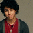 Pixelated portrait of a person with dark curly hair wearing red sleeveless top over gray long sleeves against neutral background.