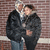 Two people stand close together against a brick wall background, with one person's arm around the other in a static pose.