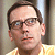Static pixelated image of a person with short brown hair, glasses, and light skin wearing a yellow shirt against a blurred background.