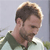 A pixelated close-up of a person with short brown hair and a beard, wearing a dark green shirt against a light gray background.