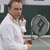 A person in a white shirt holds a large gray circular object with a dark center against a dark background.