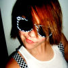 A person with short brown hair wearing a white tank top and black-and-white checkered straps, holding sunglasses near their face.