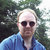 Pixelated image of a person with light hair wearing dark sunglasses and a blue shirt against a blurred green background.