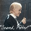 Two characters in dark robes with white collars face each other against a stone wall background, one holding a wand and the other gesturing.